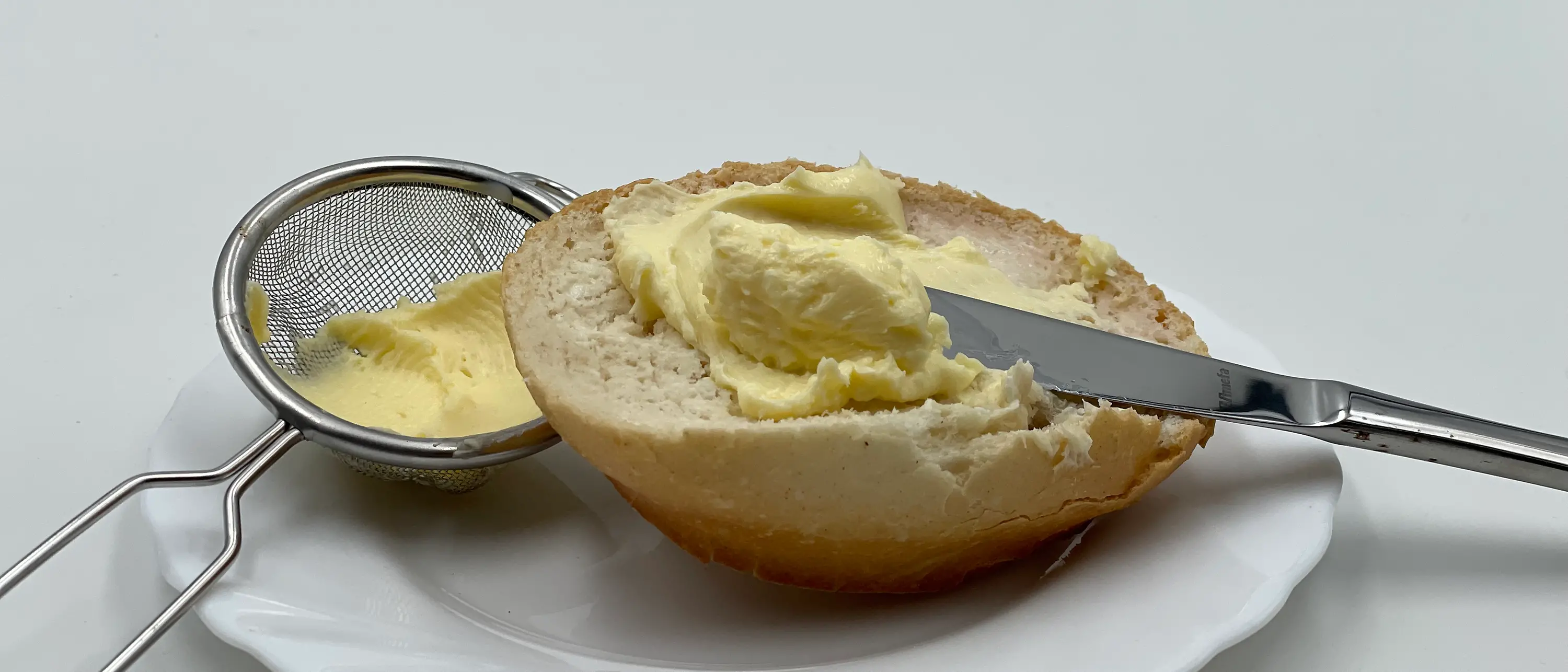 Ein halbes Brötchen mit Messer und Butter auf einem weißen Teller neben einem kleinen Sieb mit Butter