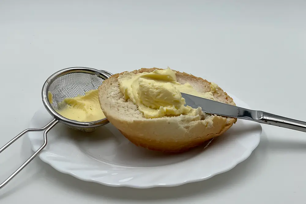 Buttered bun with knife on a plate next to a sieve with butter