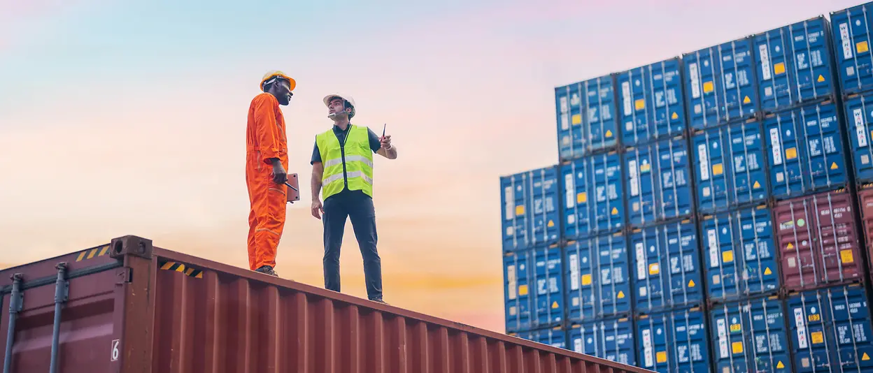 Zwei Männer stehen auf einem Container in einem Handelshafen und sprechen miteinander.